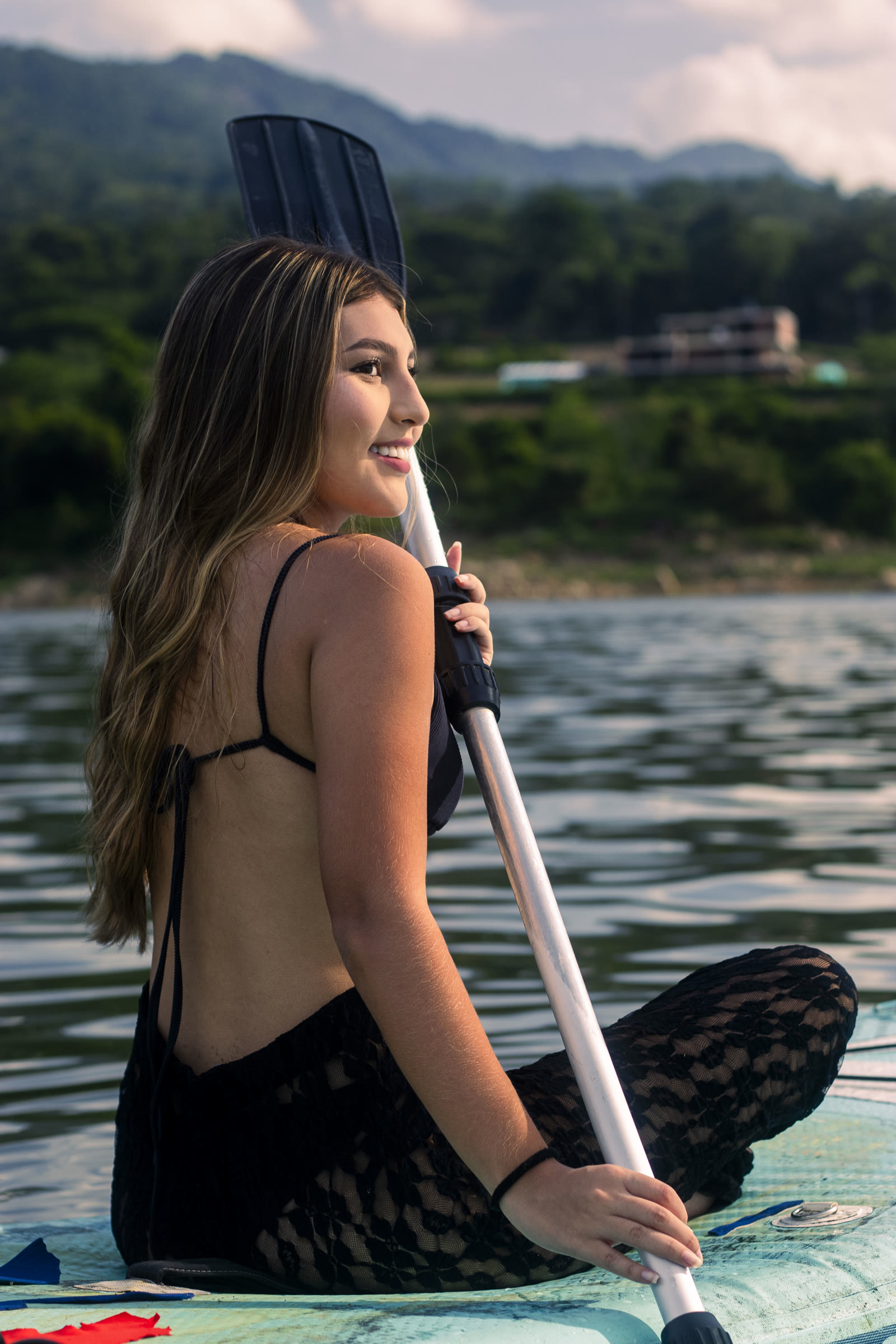 Mujer sonriendo sentada sobre una tabla de paddleboard en el Embalse de Topocoro, con montañas verdes al fondo