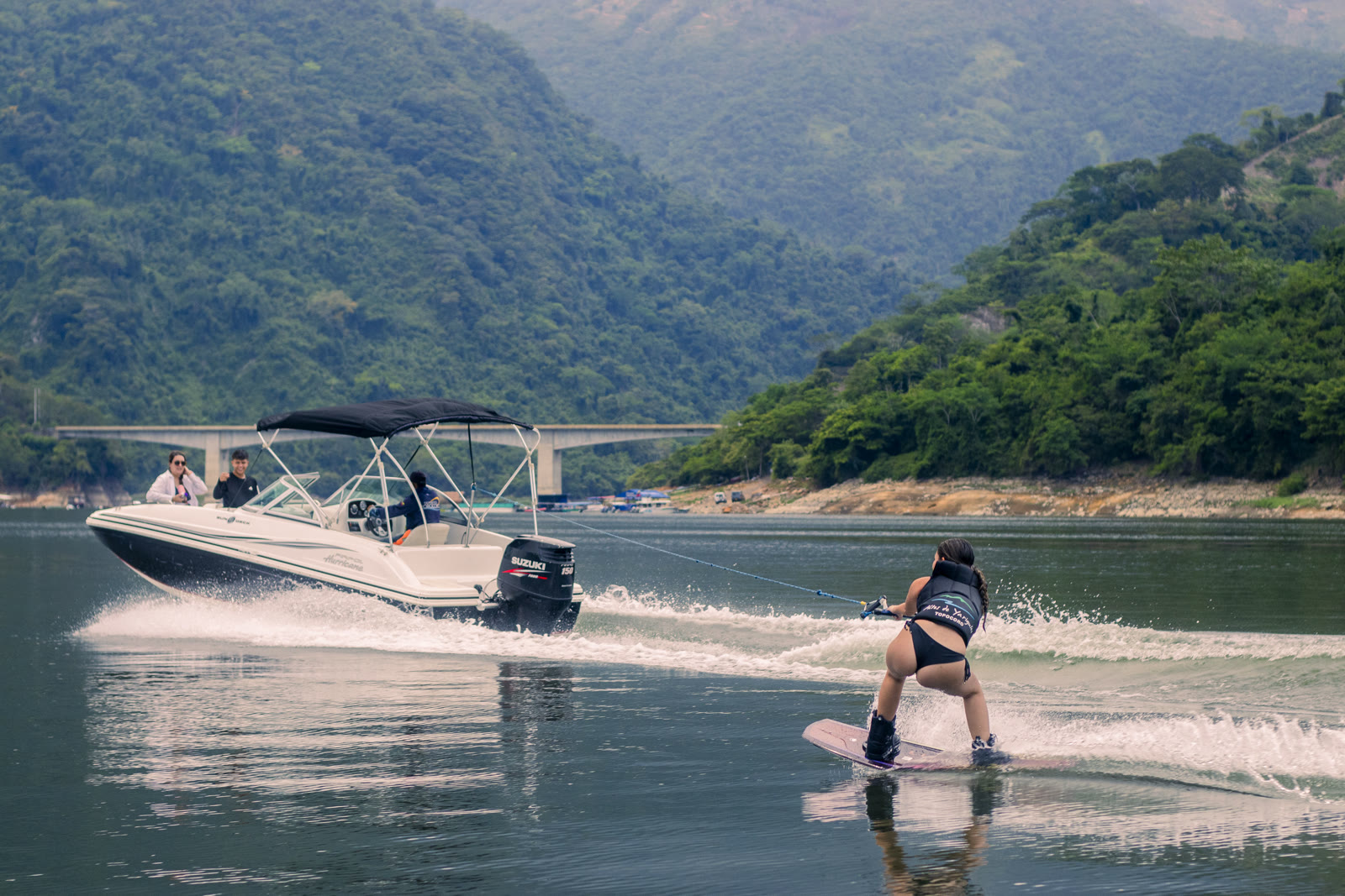 Una mujer practica wakeboard detrás de una lancha sobre el Embalse de Topocoro, con montañas verdes y un puente al fondo